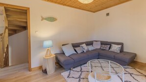Un salon chaleureux avec un canapé d’angle gris, une petite table d’appoint en bois surmontée d’une lampe, une table basse en verre et un escalier avec des rampes en bois.