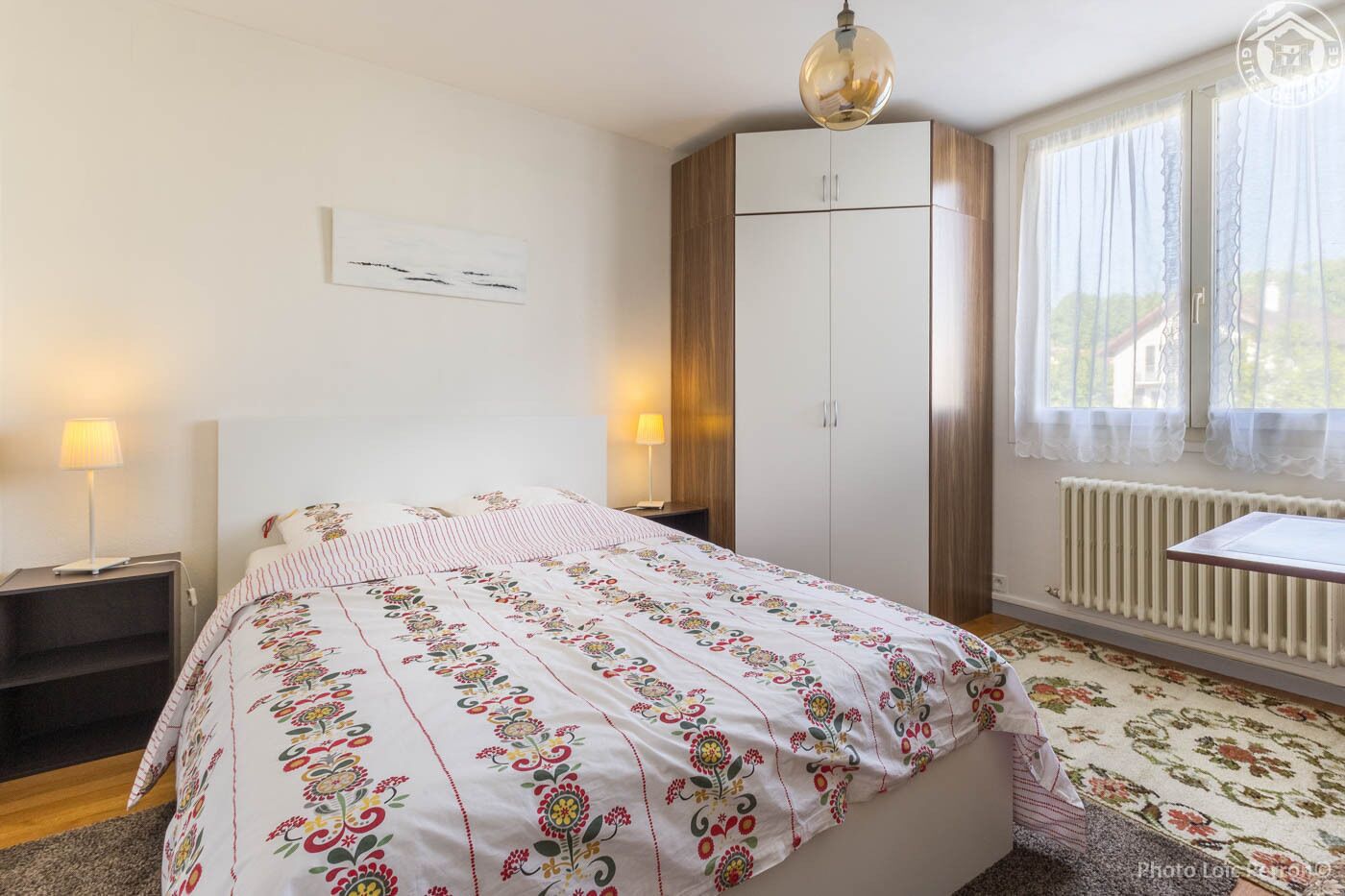 Un dormitorio con una cama, dos lámparas de noche, un armario, un radiador y una ventana con cortinas.