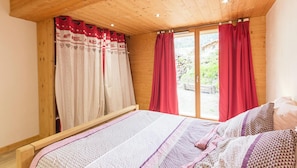 A bedroom with a wooden ceiling, a bed with patterned bedding, a window with red curtains, and a view of a garden.
