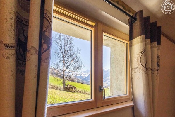 Una ventana con vista a un árbol y montañas, cortinas con un diseño, y un marco de madera.