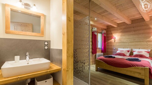 Une salle de bain moderne avec un grand lavabo, un miroir et des étagères en bois. La chambre est dotée d’un lit en bois avec une literie rouge et offre une vue sur l’extérieur grâce à une fenêtre ornée de rideaux rouges.