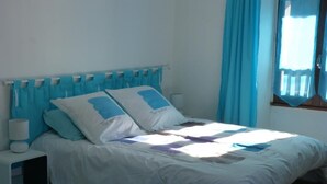 A bed with a white and blue bedspread, a nightstand with a lamp, and a window with blue curtains. - Chez Desire - St-pierre-d'entremont (SAINT PIERRE D'ENTREMONT)