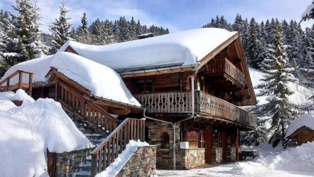 Un chalet en bois avec un escalier en pierre et un balcon, entouré d’arbres couverts de neige.