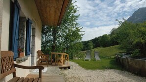 Una terraza de madera con un banco y una mesa, un muro de piedra con contraventanas azules, y vistas a una colina cubierta de hierba con árboles y una montaña al fondo.