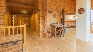 Interior de una cabaña de troncos con una escalera de madera, una pequeña mesa con sillas y una estantería.