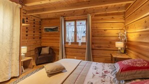 Un dormitorio de cabaña rústica con cama, silla, mesita de noche y ventana con cortinas.