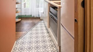 Eine schmale Küche mit einem gemusterten Boden, einem Kühlschrank und einem Fenster mit durchsichtigen Vorhängen.