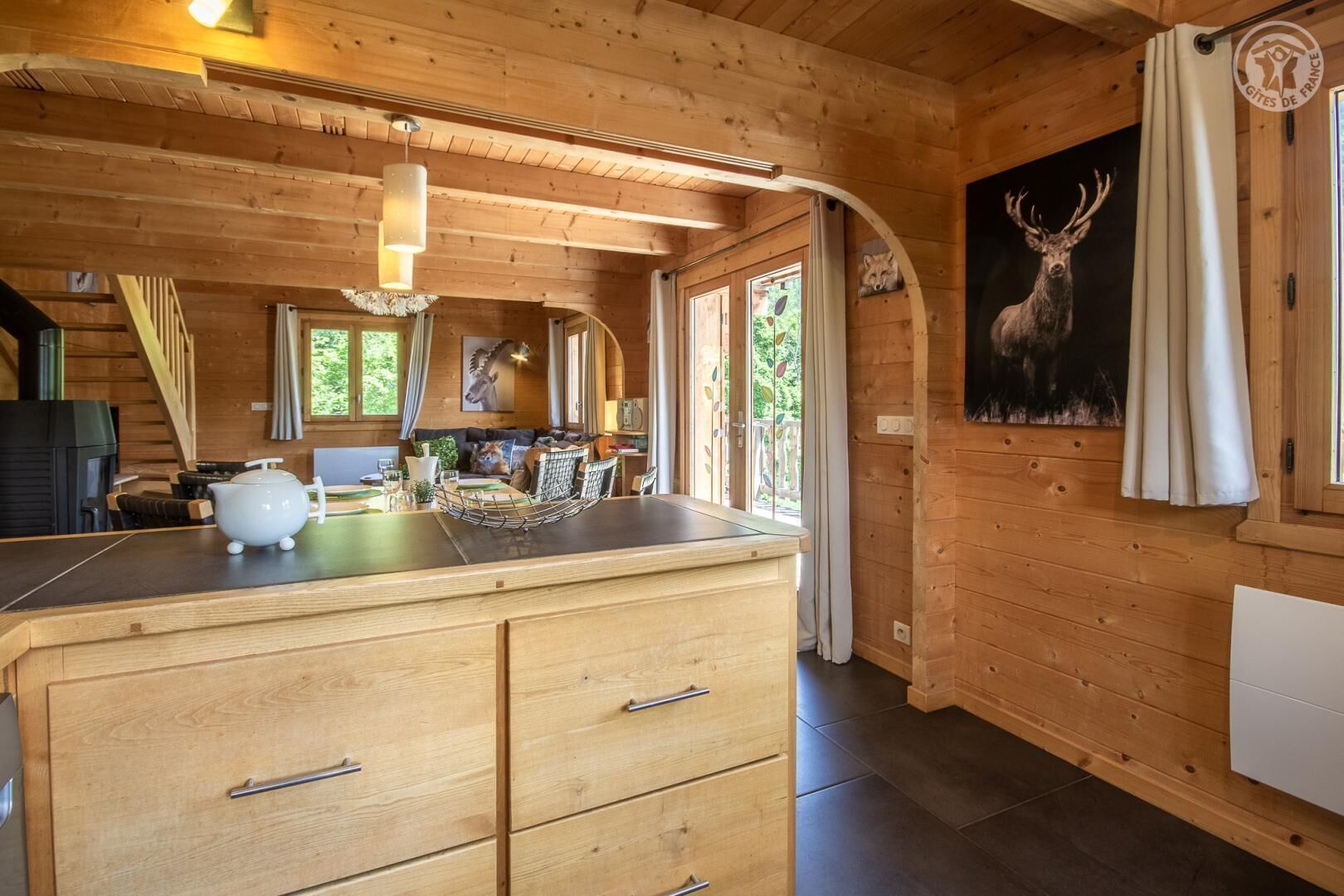 L’intérieur chaleureux d’un chalet, avec un revêtement mural en bois, un coin repas comprenant une table et des chaises, une cuisine dotée d’un grand meuble en bois et un tableau représentant un cerf accroché au mur.