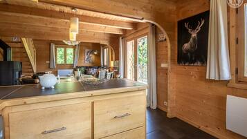 L’intérieur chaleureux d’un chalet, avec un revêtement mural en bois, un coin repas comprenant une table et des chaises, une cuisine dotée d’un grand meuble en bois et un tableau représentant un cerf accroché au mur.
