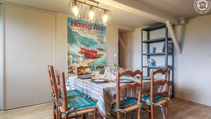 A dining area with a table set for four, surrounded by wooden chairs with blue cushions. A poster of 'Navig' Aix hangs on the wall, and a bookshelf is visible to the right. - LES Cachouds - LE Bourget-du-lac (LE BOURGET DU LAC)