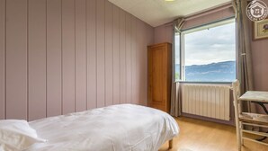 A bedroom with a bed, a wooden wardrobe, a radiator, a window with a view of mountains, and a small table with a chair. - LES Cachouds - LE Bourget-du-lac (LE BOURGET DU LAC)