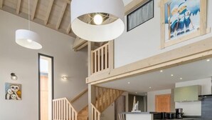 Un'ampia sala da pranzo con soffitto alto e travi a vista, una scala che conduce a un piano superiore e una zona cucina visibile sullo sfondo.