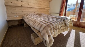 Une pièce avec un plafond en pente en bois, un lit recouvert d’une couverture en fourrure, un radiateur et une vue sur l’extérieur, visible à travers une porte en verre.