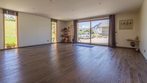 A spacious room with wooden flooring, a large sliding glass door leading to a mountain view, and a small shelf with decorative items. - LA Ferme DU Soleil - Combles (ALBIEZ MONTROND)