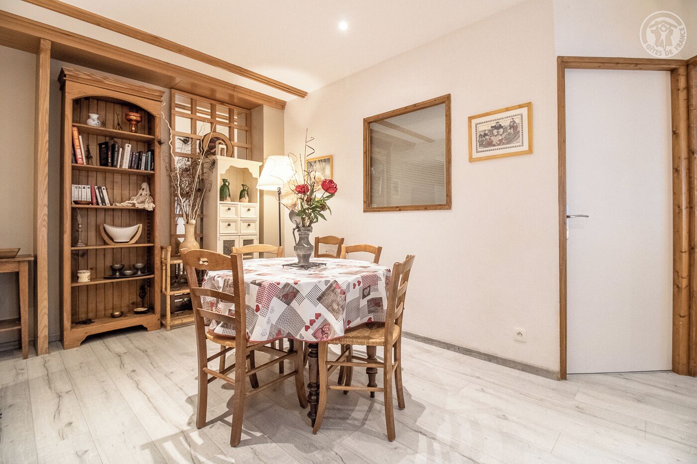 Un comedor con una mesa dispuesta para cuatro personas, rodeada de sillas de madera. A la izquierda, una estantería llena de libros y objetos decorativos. Un jarrón con flores está sobre la mesa, y un cuadro enmarcado cuelga en la pared.