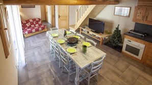 A dining area with a wooden table set for six, surrounded by white chairs. A television is on a wooden stand to the right, and a small Christmas tree is visible in the corner. - Chalet DES Alpes - Val-cenis (VAL CENIS)