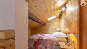Een gezellig slaapkamer op zolder met een houten plafond, een bed met sprei, een bedlampje en een klein houten tafeltje.