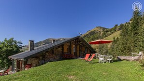 Een rustieke blokhut met schuin dak, schoorsteen en een klein terras. Er staan zitgedeeltes buiten, met een tafel en stoelen, en een rode parasol.