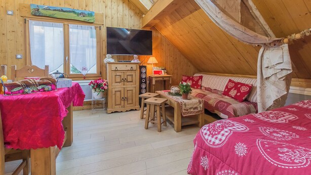 A cozy, wooden attic room with a large TV, a dining table with a red tablecloth, a sofa with red cushions, and a window with a view of the outdoors. - LA Grange Neuve - Frontenex (FRONTENEX)