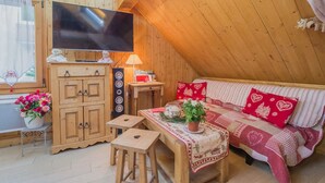 A cozy attic room with a wooden ceiling, a flat-screen TV on a cabinet, a sofa with red heart-patterned pillows, a small wooden table with a tablecloth, and a window with curtains. - LA Grange Neuve - Frontenex (FRONTENEX)