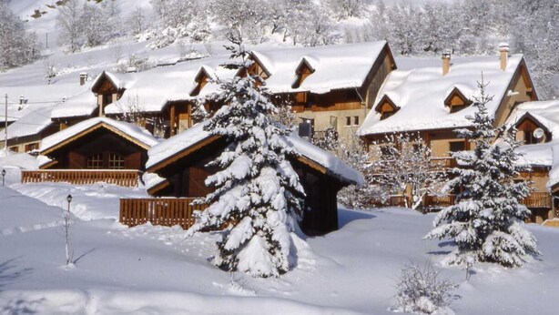 Una cabaña nevada con varias cabañas de madera, rodeada de árboles cubiertos de nieve y un paisaje montañoso.