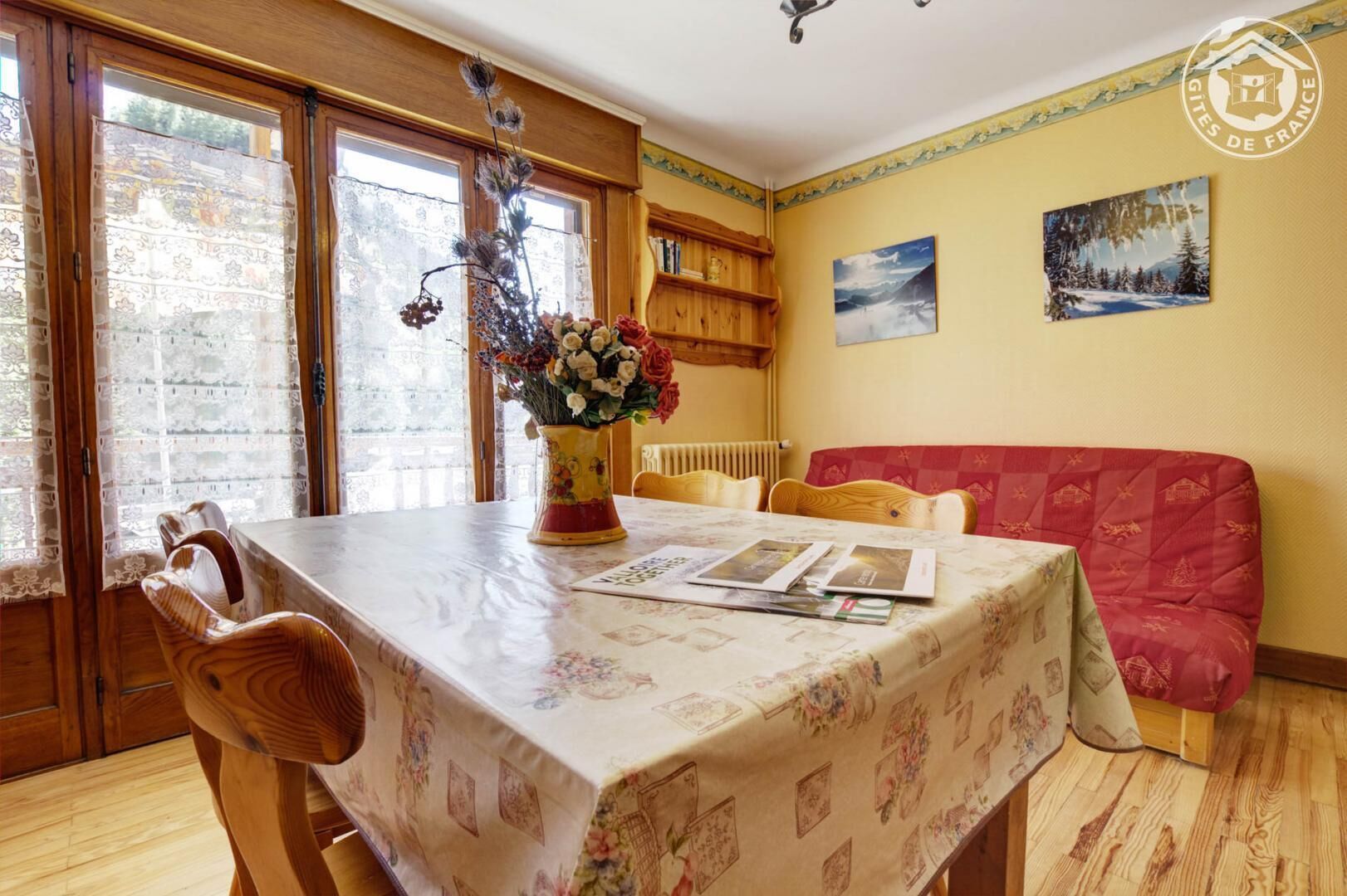 Un comedor acogedor con una mesa de madera, sillas y un florero. Se observa un sofá rojo y una estantería al fondo.