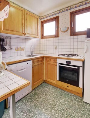 Une cuisine compacte avec des meubles en bois, un plan de travail blanc, un lave-vaisselle, un four et une fenêtre agrémentée d’un miroir décoratif.