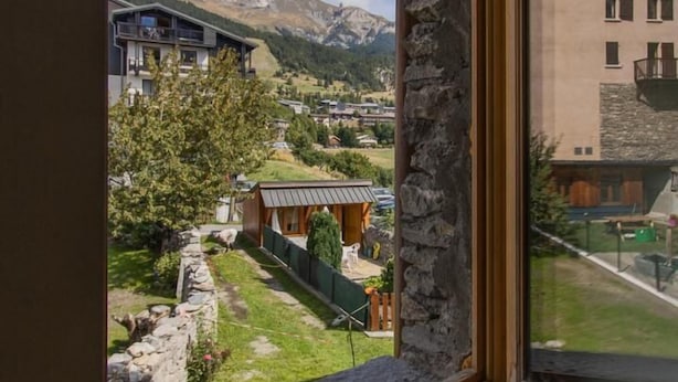 A window with a view of a mountainous landscape, stone wall, and a building with a balcony.