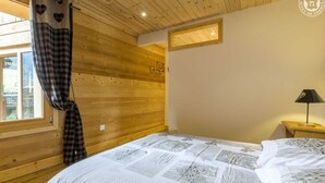 Un dormitorio con techo de madera, una cama con colcha estampada, una mesita de noche con lámpara y una ventana con cortinas.