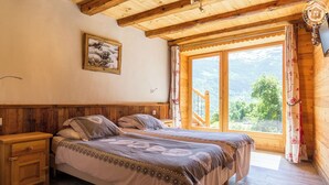 A wooden bedroom with a bed, nightstands, a painting, and a large window with a view of greenery. - Chalet Villaroger - LA Perle DES Neiges (VILLAROGER)