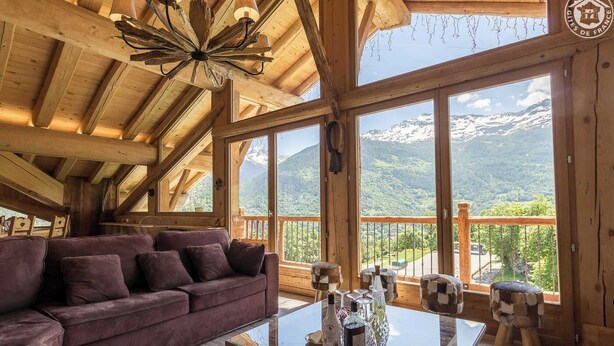 A wooden cabin with a high ceiling, a purple sofa, a glass table with bottles, and a view of snow-capped mountains. - Chalet Villaroger - LA Perle DES Neiges (VILLAROGER)