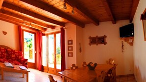 Une pièce chaleureuse avec un plafond en bois, un canapé recouvert d’un tissu à motifs de cœurs, une table à manger avec des chaises et un téléviseur fixé au mur.
