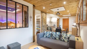 Ein modernes Wohnzimmer mit einer Couch, einem Hocker und einem runden Tisch. Im Hintergrund ist eine Küche mit Essbereich zu sehen.