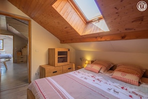 A cozy bedroom with a sloped ceiling, a skylight, a wooden cabinet, a bedside table with a lamp, and a bed with patterned bedding.