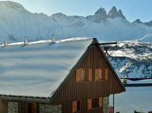 A wooden chalet with a sloped roof, stone walls, and multiple chimneys. It is surrounded by snow-covered mountains and a clear sky.