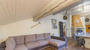 A living room with a grey sectional sofa, a wooden ceiling beam, a fireplace, and a kitchen area visible through an open doorway. - LE Boujou - Hauteluce-les-saisies (HAUTELUCE LES SAISIES)