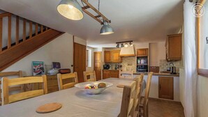 Un comedor con una mesa larga, sillas de madera y un cuenco de fruta. Hay armarios de madera, una escalera y una zona de cocina con estufa y horno.