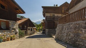 Una calle estrecha bordeada por edificios tradicionales de madera con cimientos de piedra, una pared de piedra a la derecha y una vista despejada de montañas al fondo.
