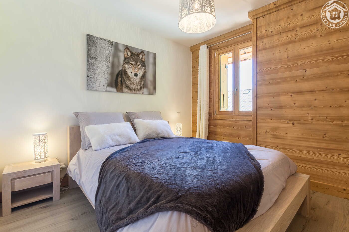 Un dormitorio con cabecera de madera, una cama grande con manta oscura, dos mesitas de noche con lámparas y un cuadro colgado en la pared con un lobo.