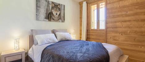 Un dormitorio con cabecera de madera, una cama grande con manta oscura, dos mesitas de noche con lámparas y un cuadro colgado en la pared con un lobo.