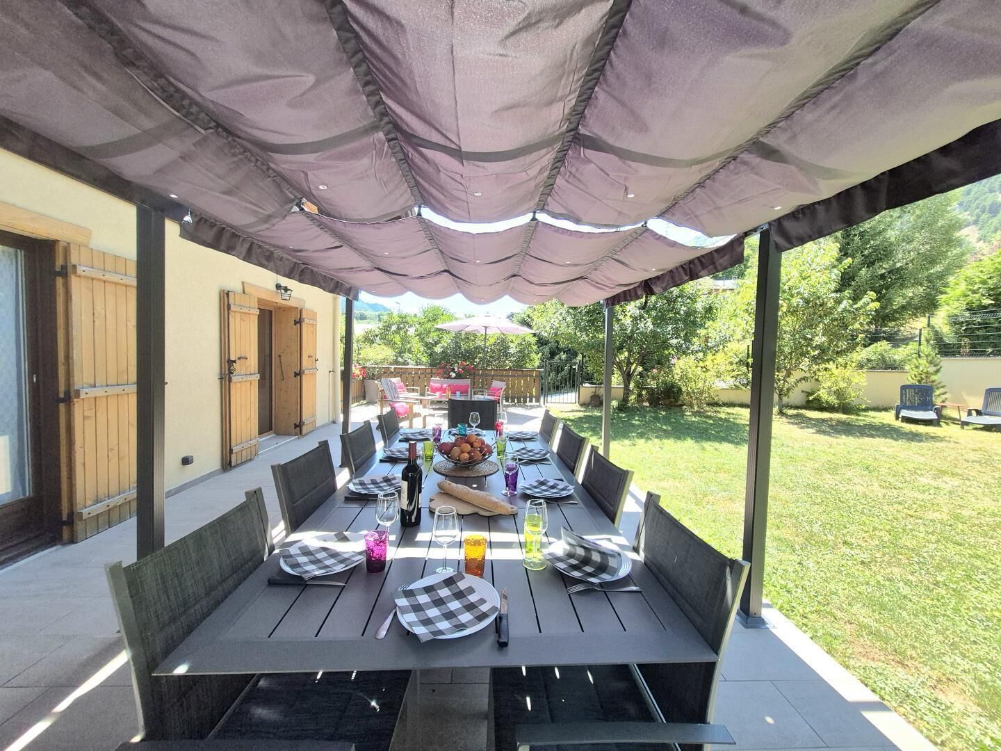 An outdoor patio with a table set for dining, surrounded by chairs, under a covered area.