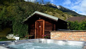 Ein Whirlpool mit einer Holzhütte im Hintergrund, umgeben von Grünpflanzen und Bergen.