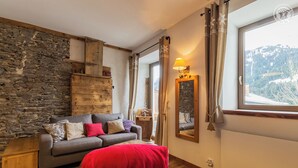 Una habitación acogedora con pared de piedra, viga de madera, sofá gris con cojines de colores, una manta roja en el suelo, un espejo y una ventana con vistas a la montaña.