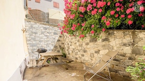 Een stenen muur met een houten tafel en een barbecue, een stoel en roze bloemen.