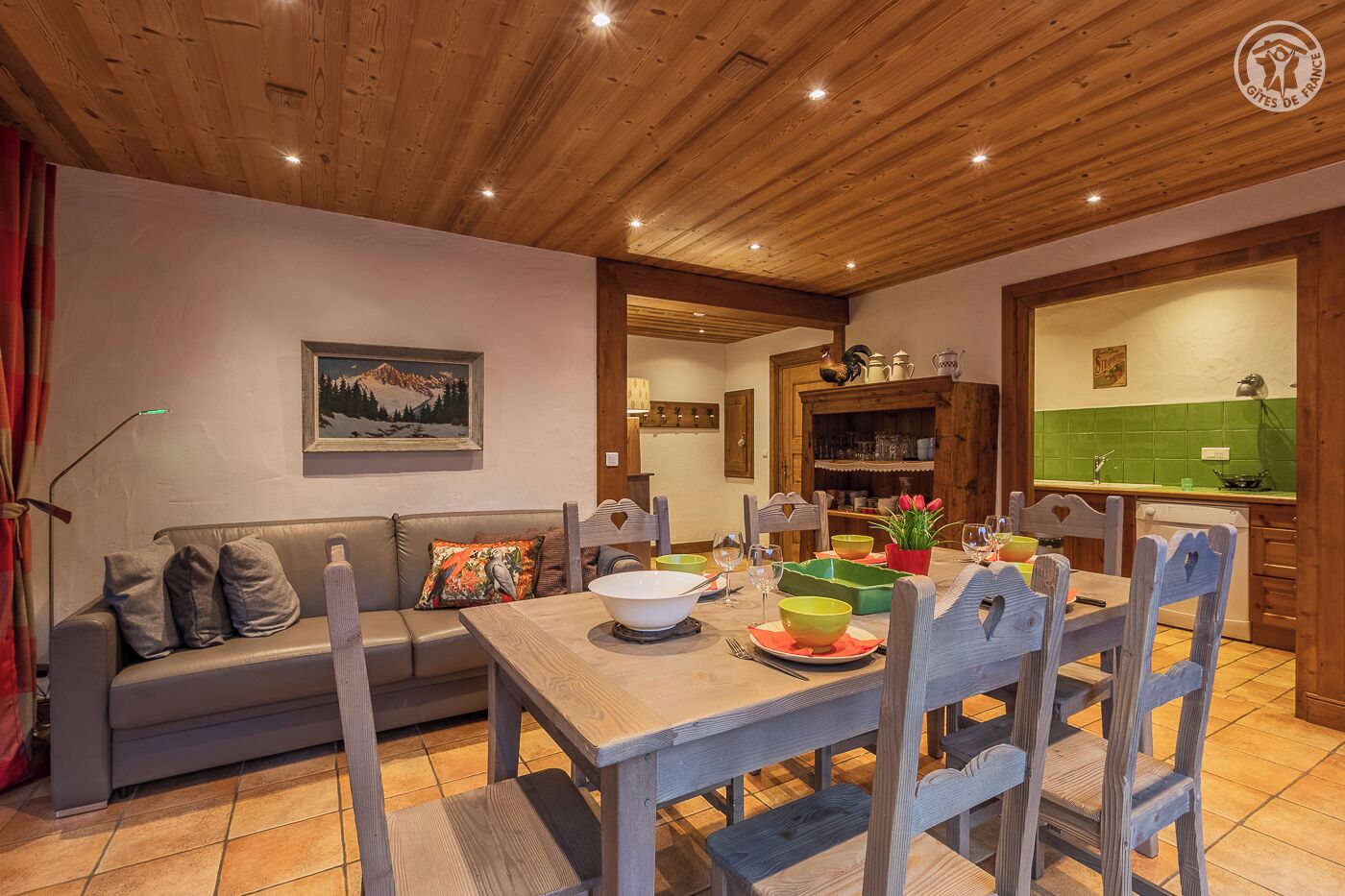 Un coin repas accueillant, avec une table en bois dressée pour quatre personnes, entourée de chaises en bois. Un canapé avec des coussins est placé contre le mur, et un tableau est accroché au-dessus.