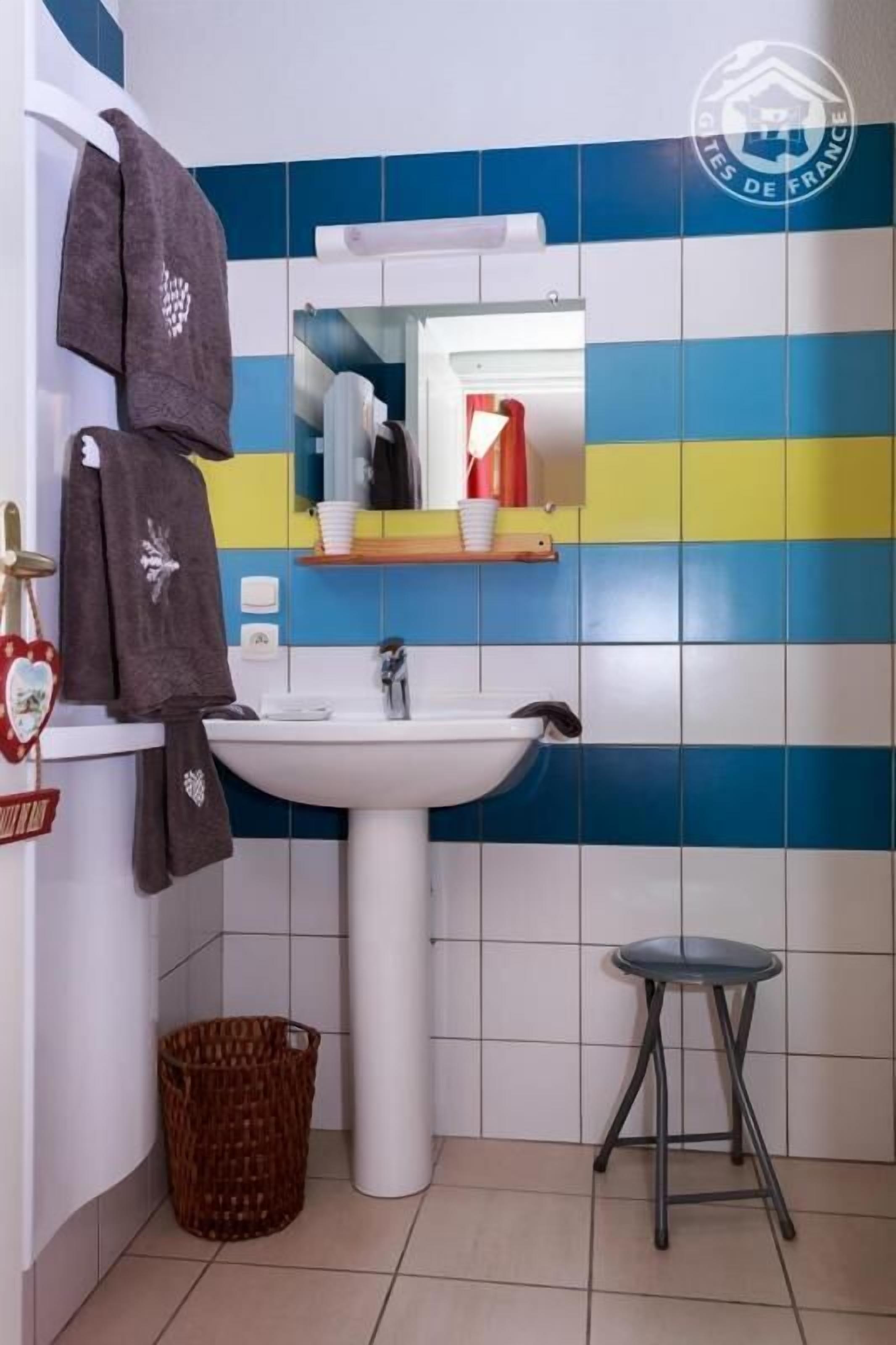 A bathroom with a white sink, a mirror, and a striped wall. There is a towel rack with towels, a wicker basket, and a stool.
