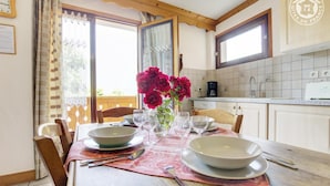 A dining area with a wooden table set for four, a vase of red flowers, and a kitchen in the background. - Gite LES Emptes - Aigueblanche (AIGUEBLANCHE)