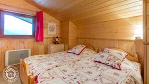 Une chambre dans un chalet en bois, avec deux lits, une fenêtre ornée d’un rideau rouge, un radiateur et une petite table sur laquelle est posé un objet décoratif.