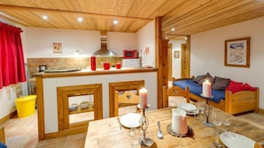 A cozy kitchen with wooden ceiling and walls, a dining area with a table set for four, and a bed with a blue blanket and red pillows. - Chalet Chaudanne - LO Soli (SAINTE FOY TARENTAISE)