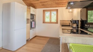 A modern kitchen with a white refrigerator, oven, and microwave, wooden ceiling, and a window with a view of greenery.